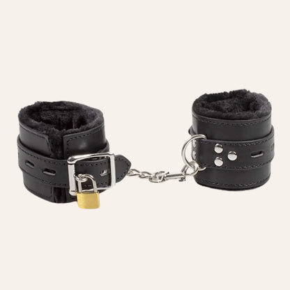 Gros plan des menottes BDSM vegan en cuir noir avec doublure en peluche et chaîne de connexion, mettant en valeur la boucle et le verrou de sécurité.