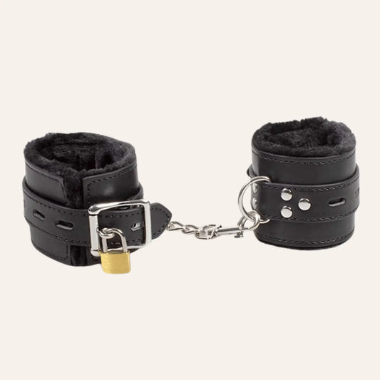 Gros plan des menottes BDSM vegan en cuir noir avec doublure en peluche et chaîne de connexion, mettant en valeur la boucle et le verrou de sécurité.