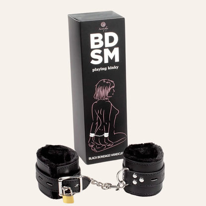 Paire de menottes en cuir noir vegan avec doublure en peluche, accompagnée de son emballage marqué "BDSM - Playing Kinky", avec une illustration minimaliste d'une personne attachée.